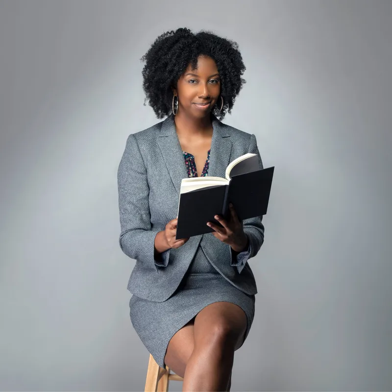 An author in a grey tweed suit sitting on a wooden stool and holding an open book, against a light grey studio backdrop