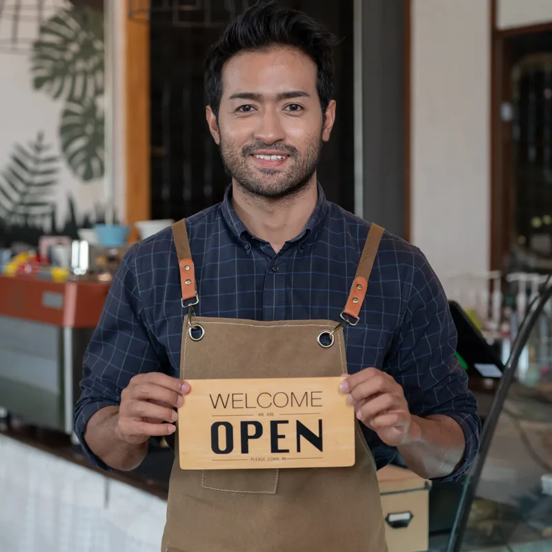 A cafe owner in a tan apron standing in his shop, smiling and holding a wooden sign reading WELCOME WE ARE OPEN