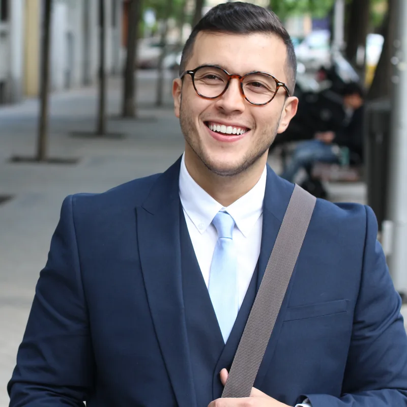 A professional in a navy three-piece suit and glasses, carrying a satchel strap and smiling on a city street